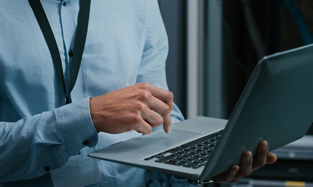 A I.T. Support worker uses the trackpad on a laptop while standing in a computer server room.