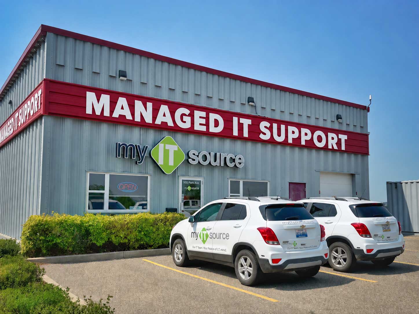 The exterior of myITsource office, a gray metal clad building with red trim and a large red sign with white letters across the entire front of the building that says "COMPUTER - SALES - SERVICE". Two white myITsource cars sit in the parking lot in front of the main entrance.