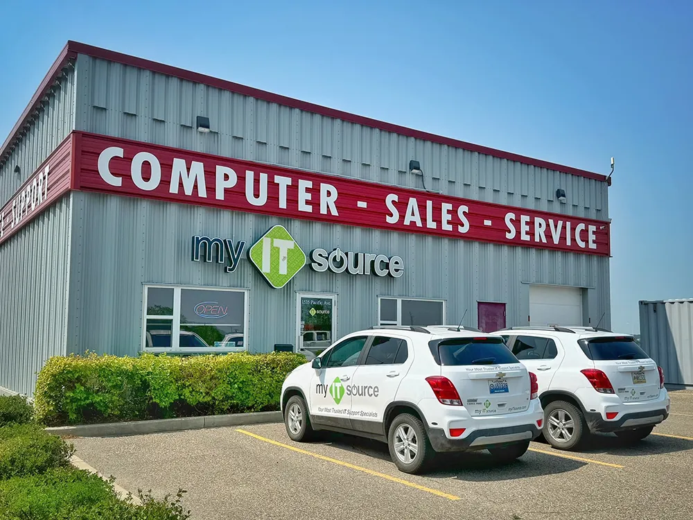 The exterior of myITsource office, a gray metal clad building with red trim and a large red sign with white letters across the entire front of the building that says "COMPUTER - SALES - SERVICE". Two white myITsource cars sit in the parking lot in front of the main entrance.